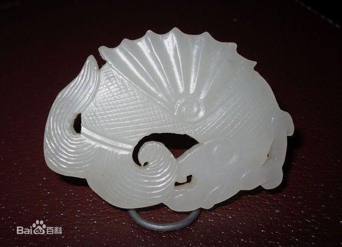 这件辽代玉摩羯鱼,长6.2厘米,高3.8厘米。为阗白玉琢制而成,局部受红褐色浸蚀。 形态为龙首鱼身,龙首发后披,龙头上出独角,鼻腔凸起,上唇上翘出尖,与上卷的下唇形成一个三角形穿孔。阴刻口角线很深,眼部呈管钻状圆眼,眉毛用细密的阴刻短线表示。龙体作鱼形,上有背鳍,胸鳍化作双翼呈展翅飞翔状,臂鳍较小用细小阴刻线表示,尾部分叉,体表刻斜网格状纹表示鱼鳞,鱼背与腹之间有一长方形穿孔可供佩戴。摩羯的形象是几种动物拼凑而成,由此可见印度的摩羯与中国的龙、凤、麒麟一样均属凭想象集中数种动物为一体的纹饰。这种纹饰大约出